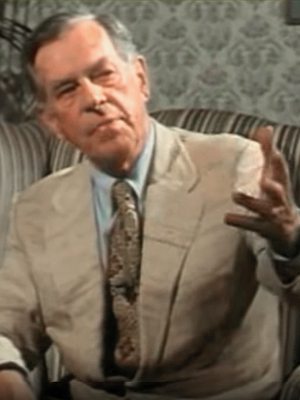 Photo of Joseph Campbell, seated, upper body, facing forward, gesturing with L hand. Video still from interview.
