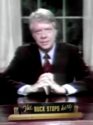 Video still, cropped, Pres. Jimmy Carter at desk, head and shoulders facing front. Desk sign says The Buck Stops Here