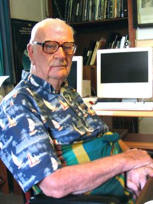 Photo of Arthur C. Clarke, old age, upper body, seated at desk in home office, facing front, slightly right