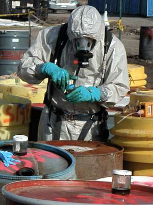 Photo of worker surrounded by metal barrels of chemical waste, in full-cover protective suit, wearing gloves and breathing mask