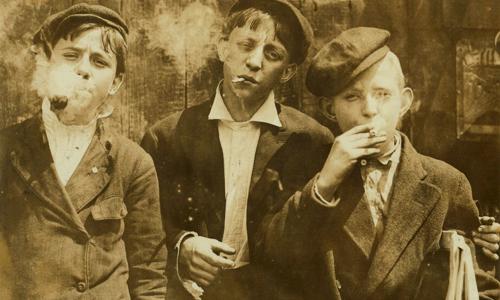 Three newsboys smoking, two with lit cigarettes in their mouths, one with a pipe puffing smoke.