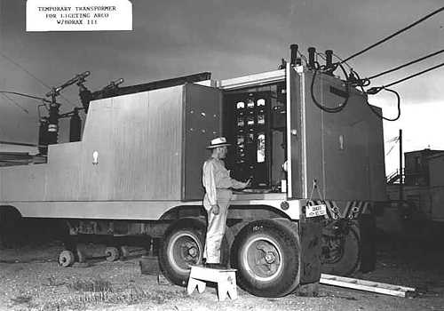 Photo of trailer-mounted mobile transformer connected to heavy electrical overhead cables. Man looks at meters on control panel