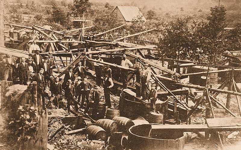 Photo of tubs and barrels collecting oil through a network of pipes. About 20 men standing among them, some in suits and hats