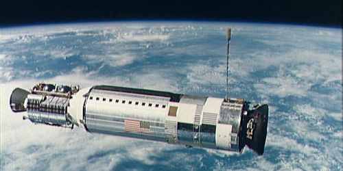 Photo in space, Agena upper stage, long cylindrical body with a squat conical docking collar at one end, earth clouds background