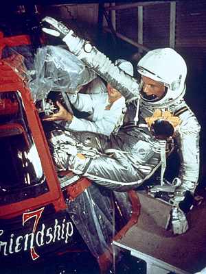 Photo of John Glenn in silver space flight suit entering his capsule feet first through a small hatch. The capsule looks small.