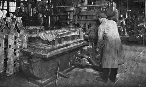 Photo of cast aluminum crank cases in a machine shop. One is on the bed of a milling machine operated by a  worker