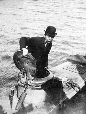 Photo of John P. Holland standing in submarine hatch, upper body revealed, hand on hatch, wearing suit and hat