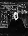 Thumbnail of Lord Kelvin at a lecture bench, with background blackboard covered with mathematical formulas