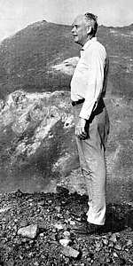 Photo of Charles Lindbergh, age 65, body full length standing on rock-strewn ground with slopes of volcano Krakatau behind him