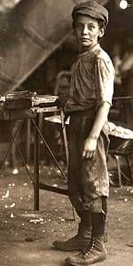 Detail from sepia photo of glass factory showing weary slim young boy, work clothes, full body standing half-left, facing front