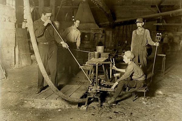 Sepia photo. At the feet of a glass blower, boy sits on stool. Each face an iron mold on floor between them. Boy holds handles.