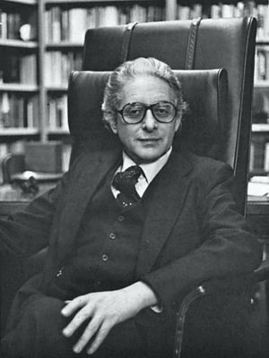 Derek J. de Solla Price, wearing glasses, upper body, seated in high-back chair with library shelves of books background