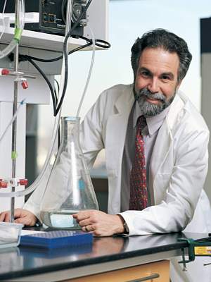 Photo of Gregg L Semenza in lab coat, behind with elbow on bench with glassware and electronic meter, upper body facing front