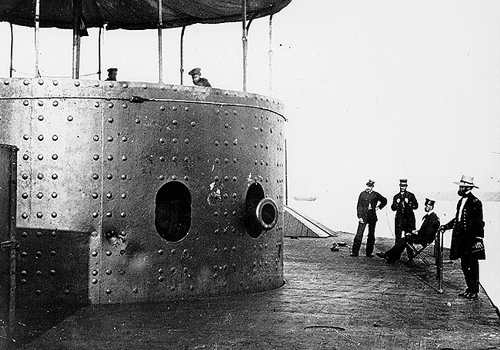 Photo showing cannon in porthole of iron plated cylindrical turret, 9 ft high, with crew standing beside it on the deck.