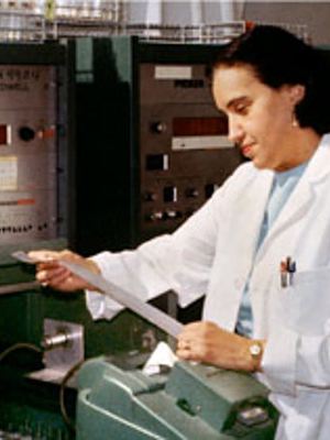 Photo of Rosalyn Yalow standing in front of lab electronic analysis equipment, upper body, facing left, in lab coat